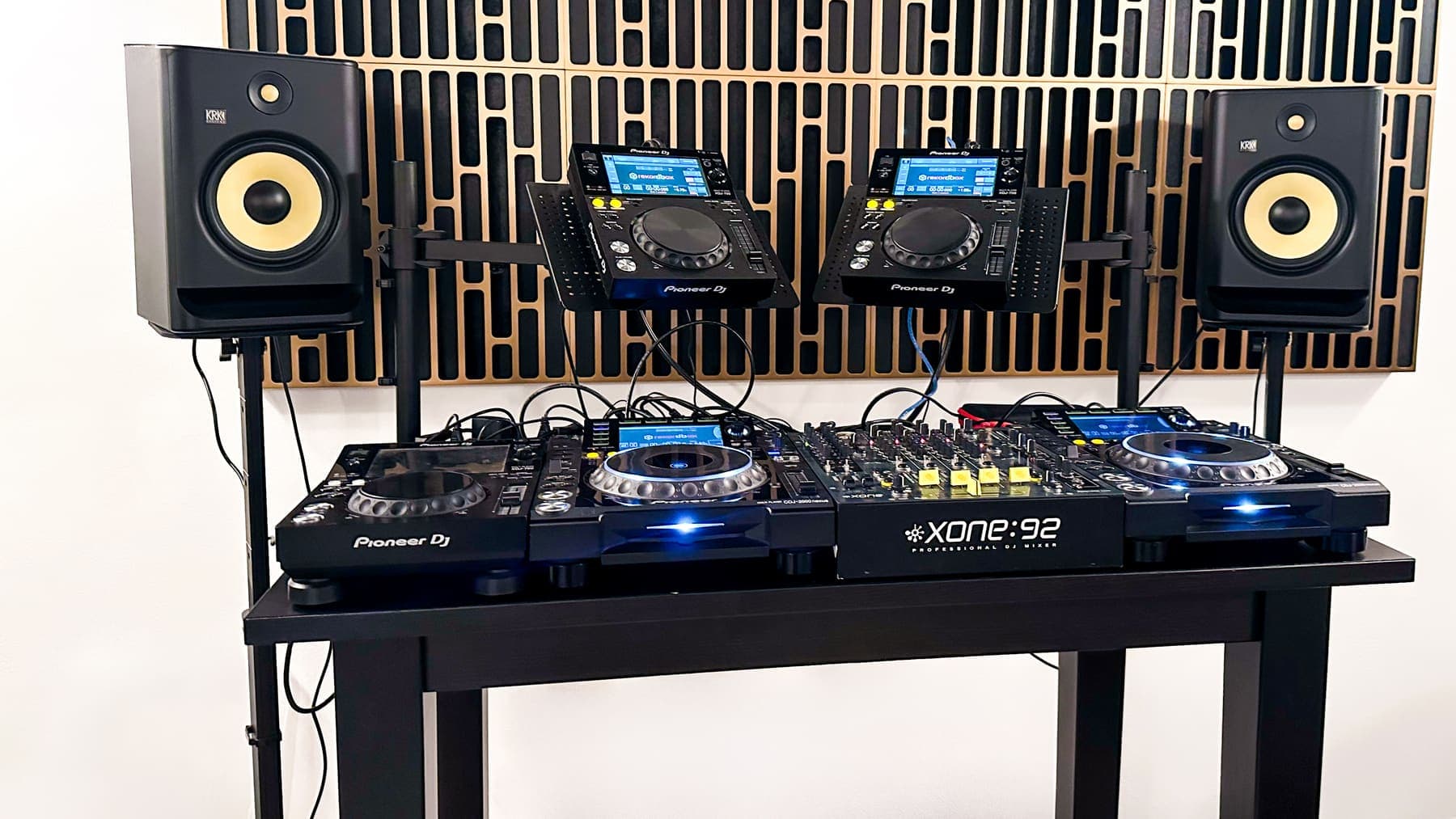 DJ-Aufbau im Studio: zwei Player und ein Mixer auf einem Tisch, dahinter zwei Studiomonitore und eine Holzlamellenwand.