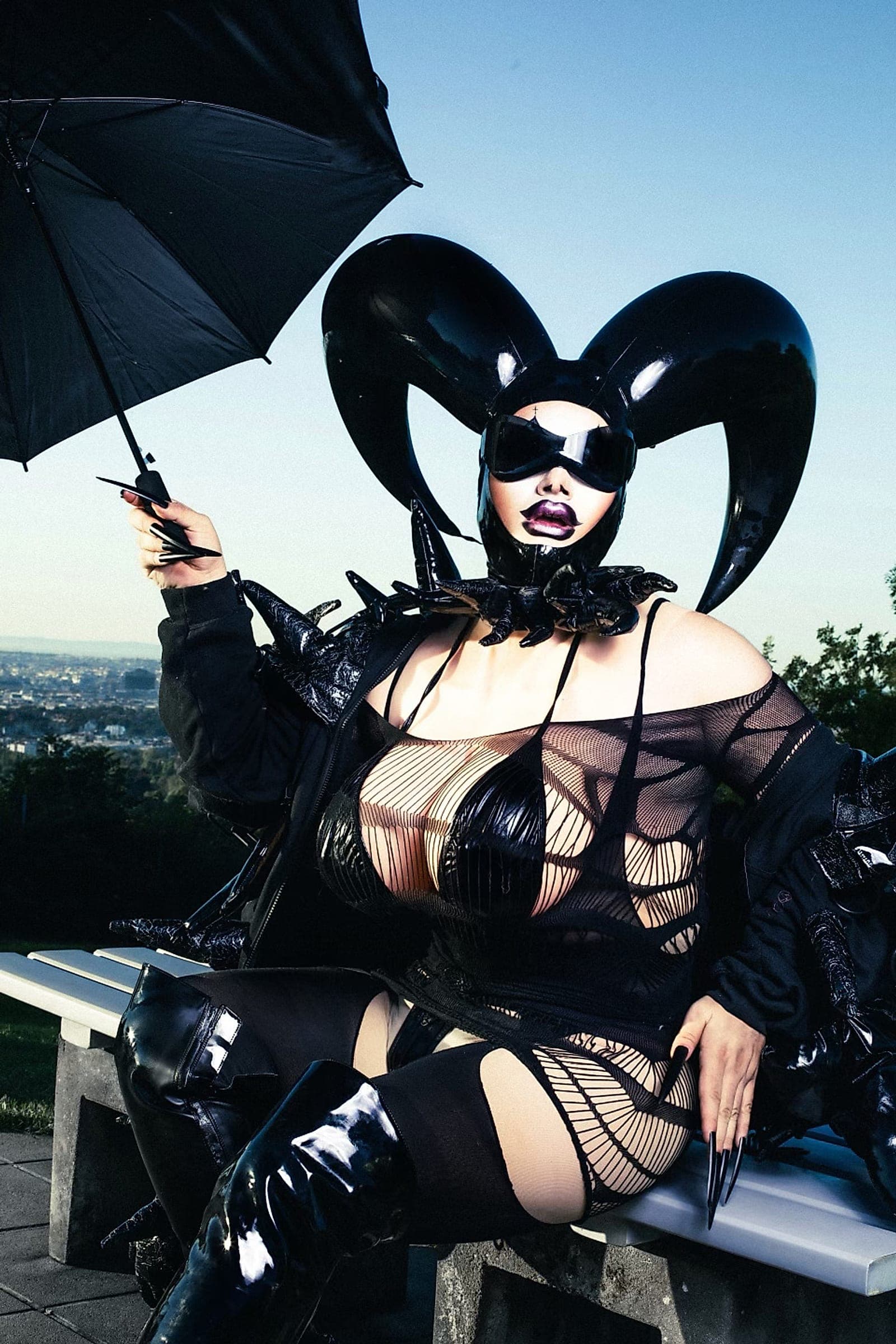Crystal O in schwarzem Latex-Outfit mit geschwungenem Kopfschmuck, einen Schirm in der Hand, vor dem Himmel über Wien.