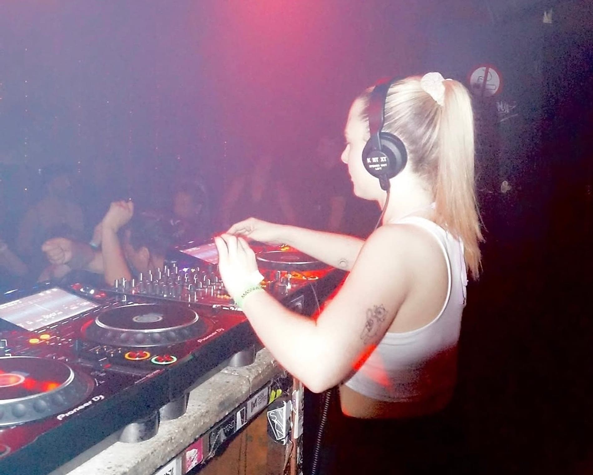 Violetta legt im Club auf, Kopfhörer auf einem Ohr, die Hände am Mischpult, dahinter das Publikum im roten Licht.