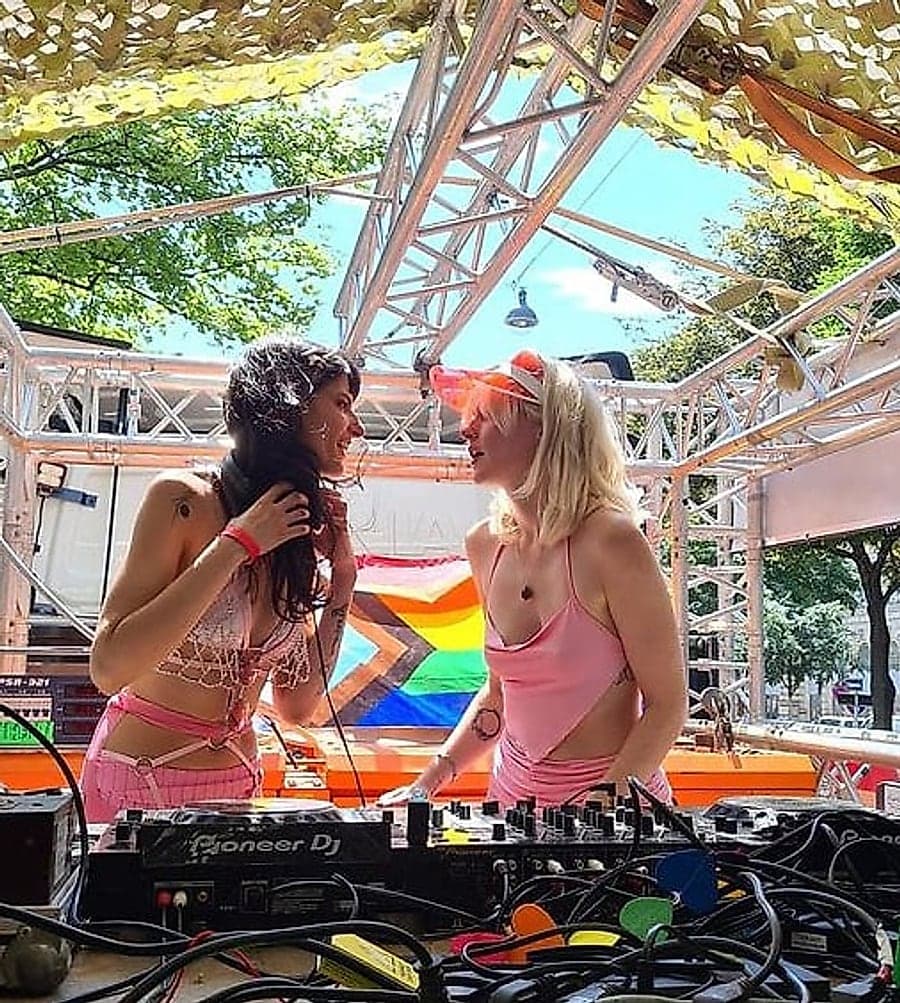 Annika Stein und Wered Fuchs gemeinsam am Mischpult bei einem Auftritt im Freien, hinter ihnen die Progress-Pride-Fahne.